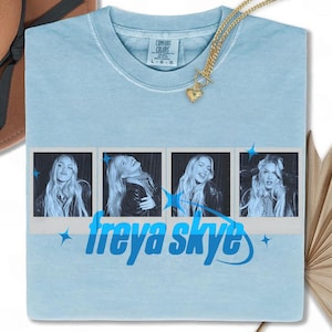 Puede incluir: Camiseta azul claro con un gráfico de cuatro fotos estilo polaroid y el texto "freya skye" en azul. Un collar dorado con un colgante de corazón está drapeado sobre la camiseta. La camiseta tiene una etiqueta Comfort Colors.