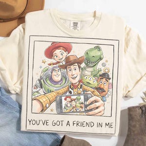 May include: Cream-colored t-shirt featuring a graphic of Toy Story characters taking a selfie. The image includes Woody, Buzz Lightyear, Jessie, Rex, and other characters. The text "YOU'VE GOT A FRIEND IN ME" is printed at the bottom.