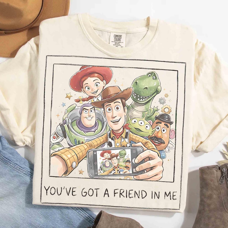 May include: Cream-colored t-shirt featuring a graphic of Toy Story characters taking a selfie. The image includes Woody, Buzz Lightyear, Jessie, Rex, and other characters. The text "YOU'VE GOT A FRIEND IN ME" is printed at the bottom.