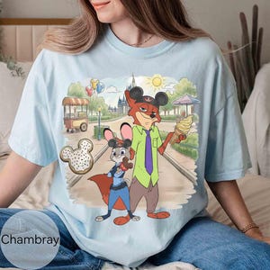 May include: Light blue t-shirt featuring a cartoon illustration of Nick Wilde and Judy Hopps from Zootopia in a park setting. Nick holds an ice cream cone, and Judy holds a bone-shaped cookie. The shirt has the word "Chambray" printed on it.