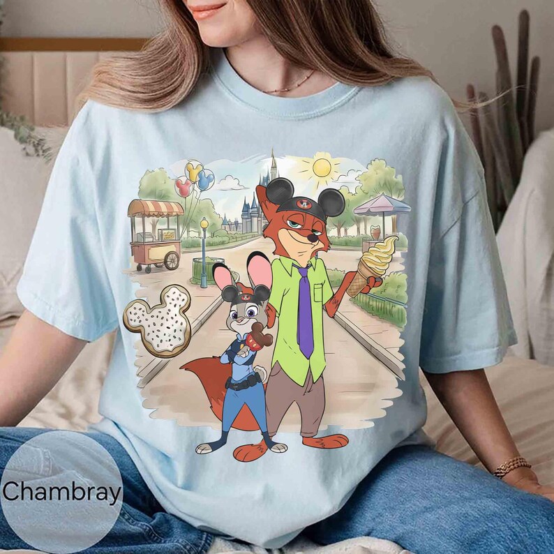 May include: Light blue t-shirt featuring a cartoon illustration of Nick Wilde and Judy Hopps from Zootopia in a park setting. Nick holds an ice cream cone, and Judy holds a bone-shaped cookie. The shirt has the word "Chambray" printed on it.