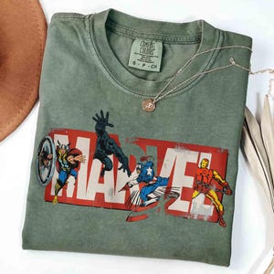 Puede incluir: Camiseta verde salvia con el logo de Marvel y ilustraciones de c&oacute;mics de Thor, Pantera Negra, Capit&aacute;n Am&eacute;rica e Iron Man. La camiseta tiene cuello redondo y mangas cortas. Un collar plateado con un colgante est&aacute; sobre la camiseta.