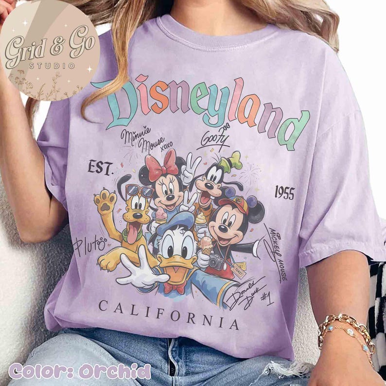 Puede incluir: Camiseta color orqu&iacute;dea con un dise&ntilde;o de Disneyland que presenta a Mickey Mouse, Minnie Mouse, Donald Duck, Goofy y Pluto. El dise&ntilde;o incluye el texto "Disneyland California 1955". La camiseta tiene un aspecto vintage ligeramente descolorido.