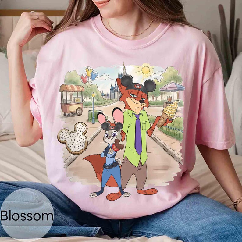 May include: A light pink t-shirt featuring a cartoon illustration of two characters from Zootopia, a fox and a rabbit, in a park setting. The fox holds an ice cream cone. A Mickey Mouse-shaped donut is in the foreground.