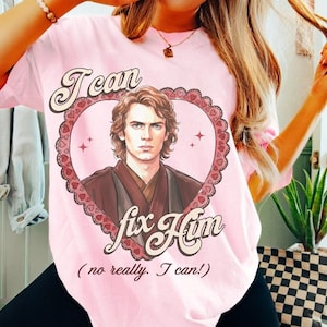 May include: Pale pink t-shirt with a heart-shaped design featuring a character portrait. The text "I can fix him" is below the portrait, with "(no really. I can!)" underneath. The design has a vintage aesthetic.