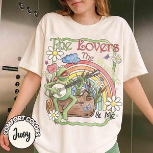 May include: A cream-colored t-shirt featuring a graphic of a green frog playing a banjo while sitting on a log. The design includes a rainbow, white daisies, butterflies, and the text "The Lovers The Dreamers & Me" in a decorative font.