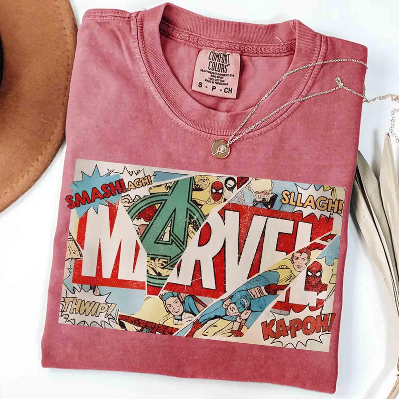 Puede incluir: Una camiseta de color rosa polvoriento con un gr&aacute;fico de c&oacute;mic de Marvel. El gr&aacute;fico incluye el logotipo de Marvel, vi&ntilde;etas de c&oacute;mic y palabras de acci&oacute;n como "SMASH!" y "KAPOW!". Un collar plateado con un colgante descansa sobre la camiseta.