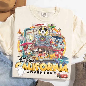 May include: Cream-colored t-shirt featuring a colorful graphic of Disney California Adventure. The design includes Mickey Mouse, a Ferris wheel, and various Disney characters. The words "Disney California Adventure" are prominently displayed.