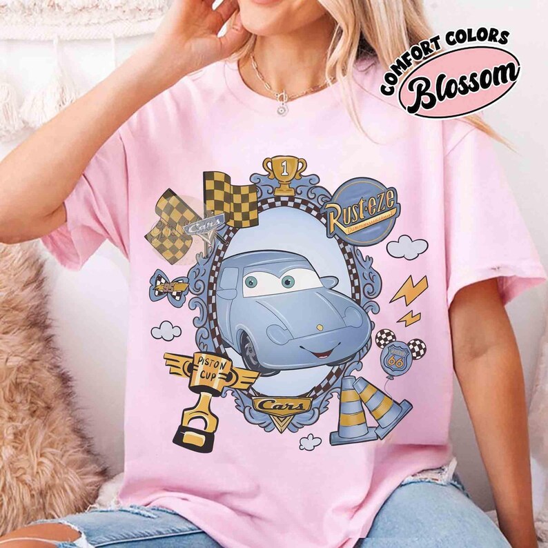 Puede incluir: Camiseta rosa claro con un dise&ntilde;o de coche de dibujos animados dentro de un marco ornamentado. El dise&ntilde;o incluye banderas de carreras, un trofeo y los textos "Rusteze" y "Cars". La camiseta tambi&eacute;n tiene el texto "Comfort Colors Blossom".