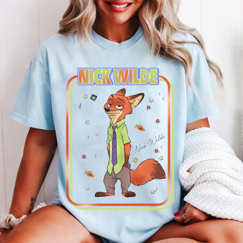 Puede incluir: Camiseta azul claro con una ilustraci&oacute;n de dibujos animados de Nick Wilde de Zootopia. El personaje lleva una camisa verde, una corbata morada y pantalones marrones. La camiseta muestra el texto "NICK WILDE" en un marco colorido, con estrellas y zanahorias.
