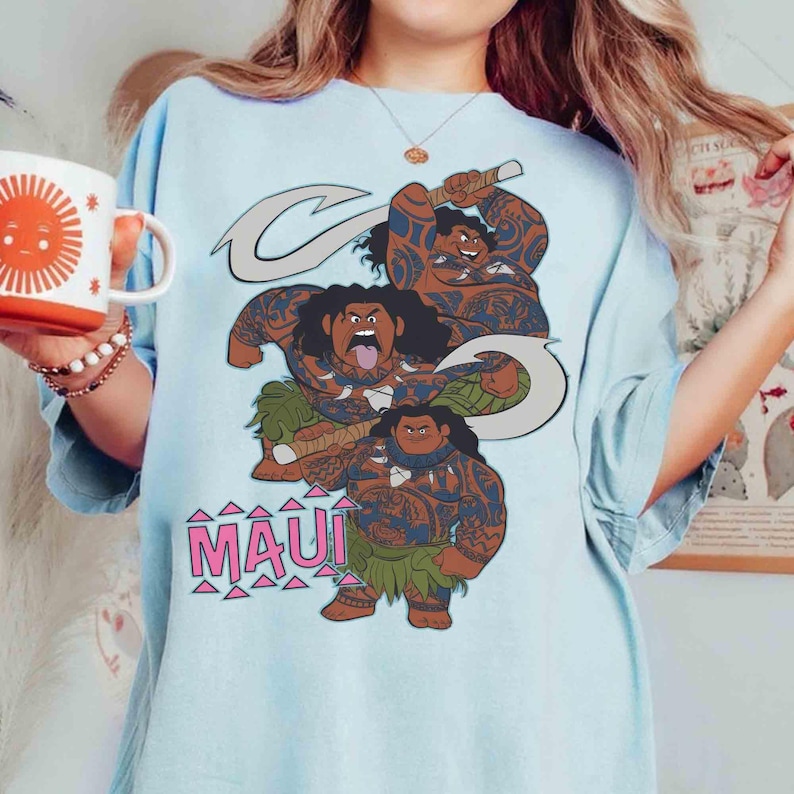 May include: Light blue t-shirt featuring a graphic of the character Maui from the animated film Moana. The design includes three figures of Maui with tattoos, a hook, and the word "MAUI" in pink letters. The shirt is a casual wear item.