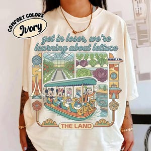Könnte beinhalten: Elfenbeinfarbenes T-Shirt mit einer farbenfrohen Grafik mit Micky Maus, Minnie Maus, Donald Duck, Daisy Duck und Goofy auf einem Boot. Der Text lautet "get in loser, we're learning about lettuce" und "The Land".