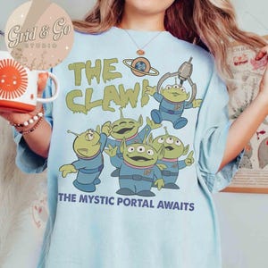 May include: Light blue t-shirt featuring a graphic of green aliens in blue uniforms with the text "THE CLAW!" and "THE MYSTIC PORTAL AWAITS." The aliens have three eyes and are in various poses. A mug with a sun design is visible.