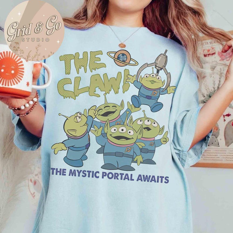 May include: Light blue t-shirt featuring a graphic of green aliens in blue uniforms with the text "THE CLAW!" and "THE MYSTIC PORTAL AWAITS." The aliens have three eyes and are in various poses. A mug with a sun design is visible.