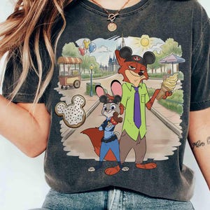 May include: Dark gray t-shirt featuring a cartoon illustration of Nick Wilde and Judy Hopps from Zootopia. Nick is wearing a green shirt, purple tie, and Mickey Mouse ears, holding an ice cream cone. Judy is in a police uniform. A Mickey Mouse-shaped donut is also shown.