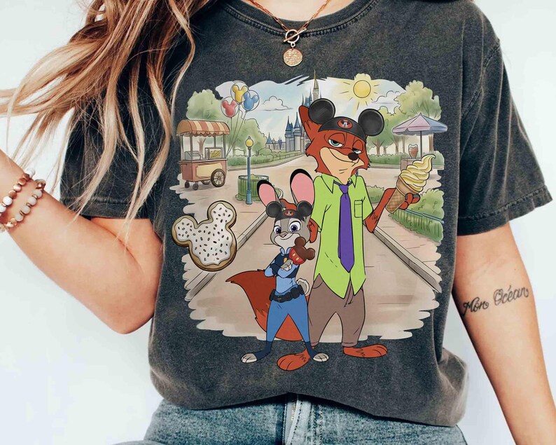May include: Dark gray t-shirt featuring a cartoon illustration of Nick Wilde and Judy Hopps from Zootopia. Nick is wearing a green shirt, purple tie, and Mickey Mouse ears, holding an ice cream cone. Judy is in a police uniform. A Mickey Mouse-shaped donut is also shown.