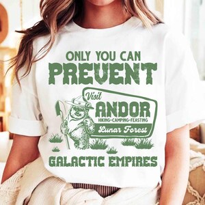 Puede incluir: Camiseta blanca con un gráfico verde. El gráfico presenta el texto "ONLY YOU CAN PREVENT" sobre un diseño con un oso sosteniendo una pala, y las palabras "Visit ANDOR" y "Lunar Forest".