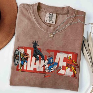 Puede incluir: Una camiseta marr&oacute;n Comfort Colors con el logo de Marvel y ilustraciones de c&oacute;mics de Thor, Black Panther, Capit&aacute;n Am&eacute;rica e Iron Man. Un collar plateado con un colgante descansa sobre la camiseta.