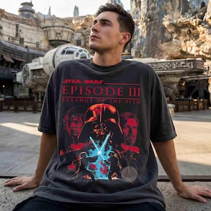 May include: A charcoal gray short-sleeve t-shirt featuring a graphic print of Star Wars Episode III Revenge of the Sith. The design includes red text, portraits of Anakin Skywalker and Obi-Wan Kenobi, and a central Darth Vader helmet.
