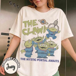 May include: Cream-colored t-shirt featuring a graphic of green aliens in blue uniforms with the text "THE CLAW!" and "THE MYSTIC PORTAL AWAITS." The design includes a planet and a claw machine. The shirt is from Comfort Colors.