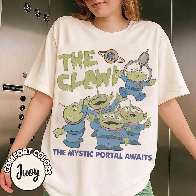 May include: Cream-colored t-shirt featuring a graphic of green aliens in blue uniforms with the text "THE CLAW!" and "THE MYSTIC PORTAL AWAITS." The design includes a planet and a claw machine. The shirt is from Comfort Colors.