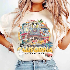 May include: Cream-colored t-shirt featuring a colorful graphic of Disney California Adventure. The design includes Mickey Mouse, a Ferris wheel, and various Disney characters. The words "Disney California Adventure" are prominently displayed.