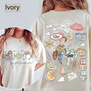 Op de afbeelding: Ivoor T-shirt met cartoonfiguren en de tekst "You got a friend in me!". Het ontwerp bevat Woody, Buzz Lightyear en andere Toy Story-elementen. Het shirt heeft een ronde hals en korte mouwen.