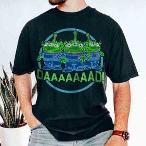 May include: Black t-shirt featuring a graphic of three green aliens with three eyes each, wearing blue jumpsuits. The aliens are inside a blue circle with the word "DAAAAAAAD!" in green below them. The shirt is a dark color.