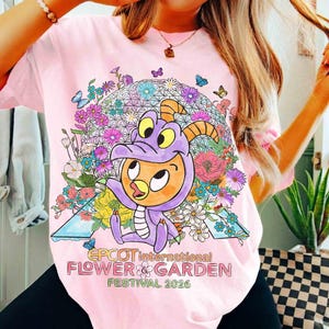 May include: Pink t-shirt featuring a colorful graphic of a cartoon dragon character in a purple costume, surrounded by flowers and butterflies. The text "EPCOT International Flower & Garden Festival 2026" is printed below the graphic.