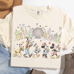 May include: Cream-colored t-shirt featuring a colorful line drawing of Disney characters, including Mickey Mouse, Minnie Mouse, Donald Duck, Goofy, and Pluto, walking in front of a stylized cityscape with the Epcot ball. The shirt has a Comfort Colors tag.