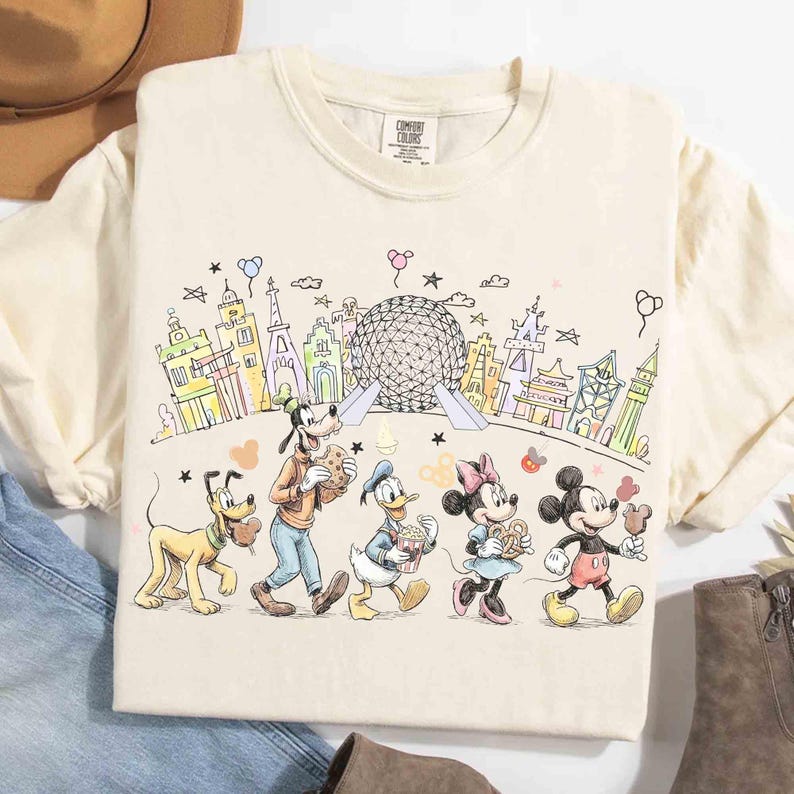 May include: Cream-colored t-shirt featuring a colorful line drawing of Disney characters, including Mickey Mouse, Minnie Mouse, Donald Duck, Goofy, and Pluto, walking in front of a stylized cityscape with the Epcot ball. The shirt has a Comfort Colors tag.