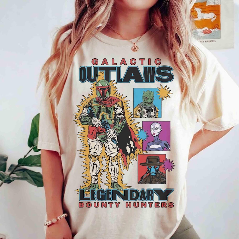 May include: Cream-coloured t-shirt with a graphic of a bounty hunter and the text "GALACTIC OUTLAWS LEGENDARY BOUNTY HUNTERS." The design includes images of various characters in a retro comic book style.