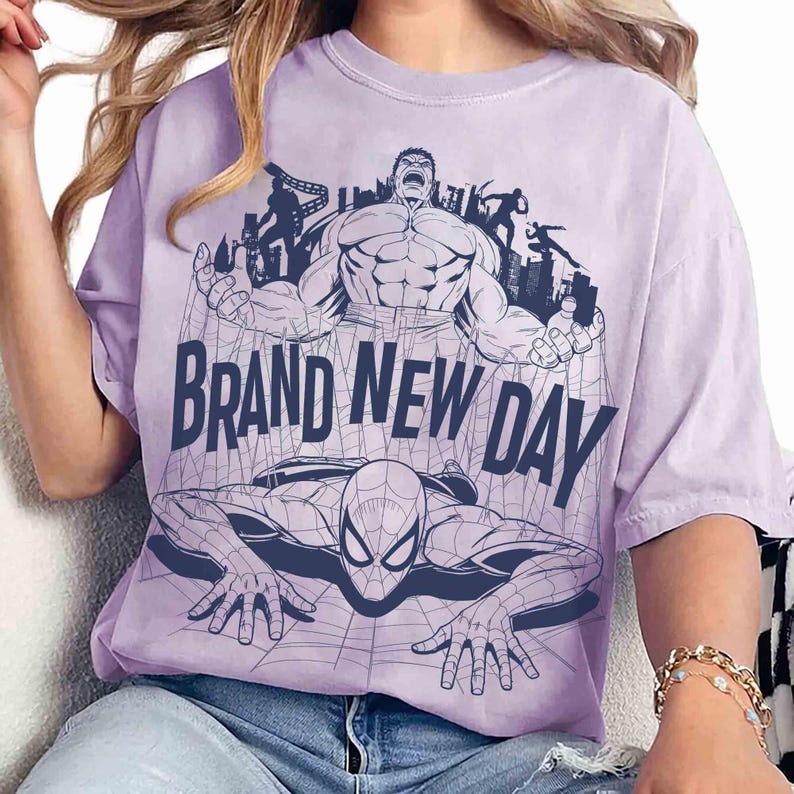 Op de afbeelding: Een lichtpaars t-shirt met korte mouwen en een donkerblauwe grafische print van de Hulk die boven Spider-Man staat. De tekst BRAND NEW DAY is in grote donkerblauwe hoofdletters in het midden van het ontwerp op het shirt gedrukt.