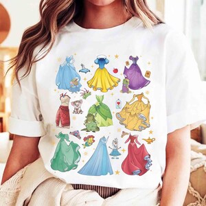 以下が含まれることがあります： 様々なプリンセスドレスのカラフルなイラストが描かれた白いTシャツ。デザインには、青、黄、緑、紫、赤の色合いのドレス、それに付随するキャラクターやアクセサリーが含まれています。このシャツはカジュアルな普段着です。