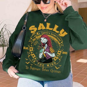 K&ouml;nnte beinhalten: Dunkelgr&uuml;nes Sweatshirt mit einem Aufdruck von Sally aus The Nightmare Before Christmas. Das Design enth&auml;lt den Text "SALLY", "I SENSE THERE'S SOMETHING", "IN THE WIND", "The Nightmare Before Christmas" und "1993".