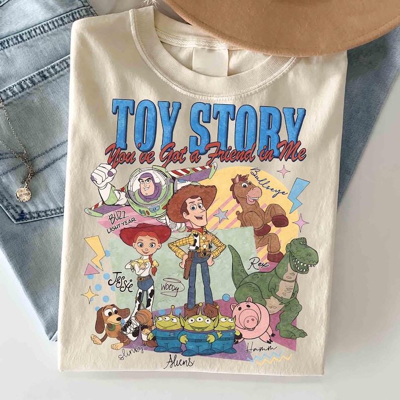 May include: Cream-colored Toy Story graphic t-shirt featuring Buzz Lightyear, Woody, Jessie, Rex, Slinky, Bullseye, Hamm, and the Aliens. The shirt has the text "TOY STORY You've Got a Friend in Me" in blue and red.