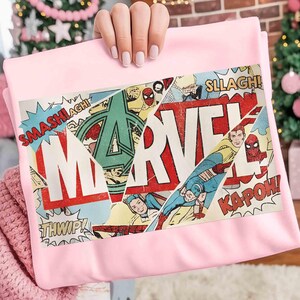 Puede incluir: Una manta rosa claro con un dise&ntilde;o de c&oacute;mic. El dise&ntilde;o presenta la palabra "MARVEL" en rojo y blanco, junto con im&aacute;genes de superh&eacute;roes y palabras de acci&oacute;n como "SMASH!" y "KAPOW!"