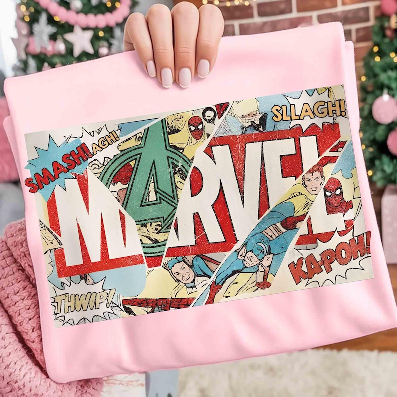 Puede incluir: Una manta rosa claro con un dise&ntilde;o de c&oacute;mic. El dise&ntilde;o presenta la palabra "MARVEL" en rojo y blanco, junto con im&aacute;genes de superh&eacute;roes y palabras de acci&oacute;n como "SMASH!" y "KAPOW!"