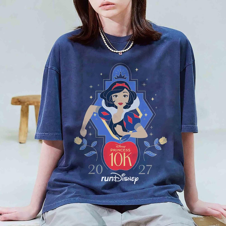 K&ouml;nnte beinhalten: Marineblaues T-Shirt mit einer Grafik von Schneewittchen vom Disney Princess 10K-Lauf. Das Design enth&auml;lt den Text "Disney Princess 10K" &uuml;ber einem roten Apfel, mit den Zahlen "20" und "27" auf beiden Seiten. Das T-Shirt hat einen Vintage-Look.
