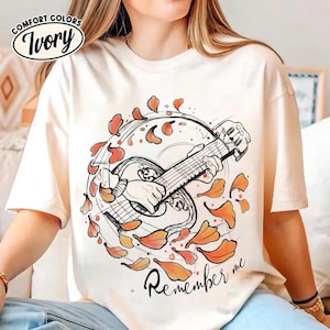 May include: Ivory-colored t-shirt with a graphic of a mandolin being played, surrounded by orange and red leaf shapes. The text "Remember me" is written in cursive below the mandolin. The shirt has the "Comfort Colors" logo.