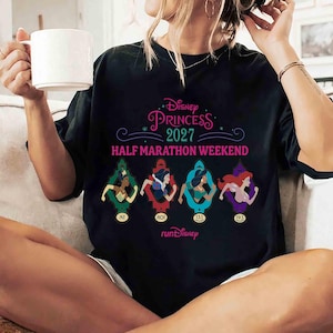 K&ouml;nnte beinhalten: Schwarzes T-Shirt mit dem Text "Disney Princess 2027 Half Marathon Weekend". Das Shirt zeigt vier Disney-Prinzessinnen-Illustrationen in farbenfrohen Rahmen, mit dem "runDisney"-Logo unten.