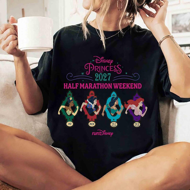 K&ouml;nnte beinhalten: Schwarzes T-Shirt mit dem Text "Disney Princess 2027 Half Marathon Weekend". Das Shirt zeigt vier Disney-Prinzessinnen-Illustrationen in farbenfrohen Rahmen, mit dem "runDisney"-Logo unten.