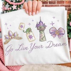 Może przedstawiać: Biała koszulka z nadrukiem przedstawiającym fioletowo-złotą tiarę, zielonego kameleona i wieżę z fioletowymi akcentami. Tekst "Go Live Your Dream" jest napisany kursywą. Projekt inspirowany jest motywem baśniowym.