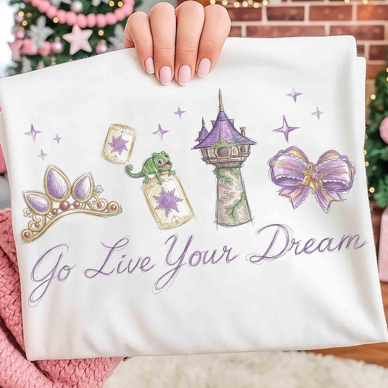 Może przedstawiać: Biała koszulka z nadrukiem przedstawiającym fioletowo-złotą tiarę, zielonego kameleona i wieżę z fioletowymi akcentami. Tekst "Go Live Your Dream" jest napisany kursywą. Projekt inspirowany jest motywem baśniowym.