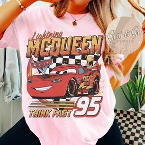 Pu&ograve; includere: T-shirt rosa chiaro con una grafica di Saetta McQueen del film Cars. Il design include i testi "Lightning McQueen", "Cars", "95" e "Think Fast". Sono visibili anche un trofeo e una bandiera a scacchi.