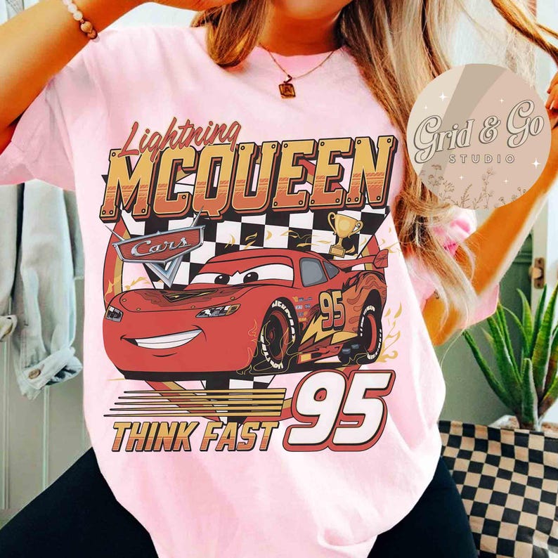 Pu&ograve; includere: T-shirt rosa chiaro con una grafica di Saetta McQueen del film Cars. Il design include i testi "Lightning McQueen", "Cars", "95" e "Think Fast". Sono visibili anche un trofeo e una bandiera a scacchi.