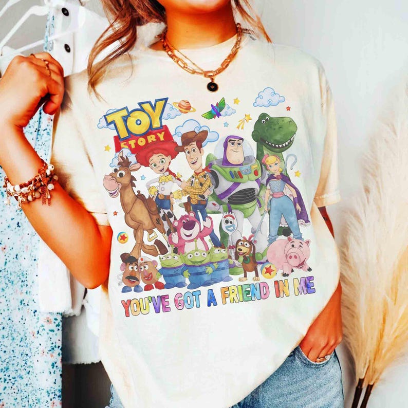 May include: Cream-coloured t-shirt with a vibrant graphic of Toy Story characters, such as Woody, Buzz Lightyear, and Rex. The text "TOY STORY" is at the top, with "YOU'VE GOT A FRIEND IN ME" below. The shirt has a relaxed fit.