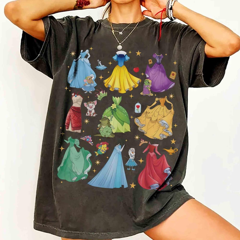 以下が含まれることがあります： 様々なプリンセスドレスのカラフルなイラストが描かれた、濃いグレーのTシャツ。デザインには、青、黄、緑、紫、赤のドレスが含まれており、キャラクターに関連する画像も添えられています。このシャツはヴィンテージ感のある外観です。