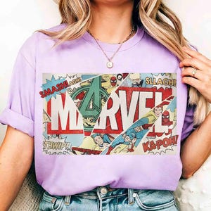 Puede incluir: Una camiseta lila claro con un dise&ntilde;o gr&aacute;fico de c&oacute;mic. La palabra "MARVEL" est&aacute; en blanco. El dise&ntilde;o incluye im&aacute;genes de superh&eacute;roes y onomatopeyas como "SMASH!" y "KAPOW!"