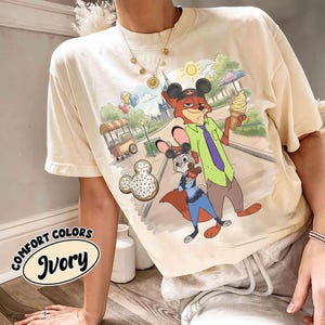May include: Ivory-colored t-shirt featuring a cartoon scene with a fox and a rabbit, both wearing Mickey Mouse ears. The fox holds an ice cream cone. The shirt also has a Mickey Mouse-shaped donut graphic and the words "COMFORT COLORS Ivory".